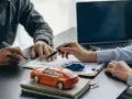 Zwei Personen unterschreiben einen Vertrag, während Modellautos und ein Laptop auf dem Tisch liegen. Dies symbolisiert den Kauf eines Autos mit Herstellergarantie.