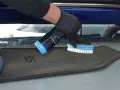 Person mit Handschuhen sprüht Reiniger auf einen grauen Autoteppich und schrubbt ihn mit einer Bürste, um ihn zu reinigen.
