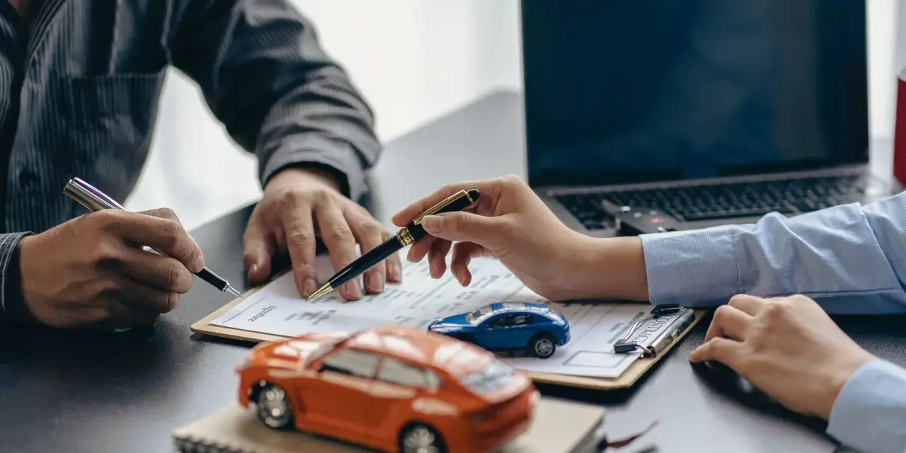 Zwei Personen unterschreiben einen Vertrag, während Modellautos und ein Laptop auf dem Tisch liegen. Dies symbolisiert den Kauf eines Autos mit Herstellergarantie.