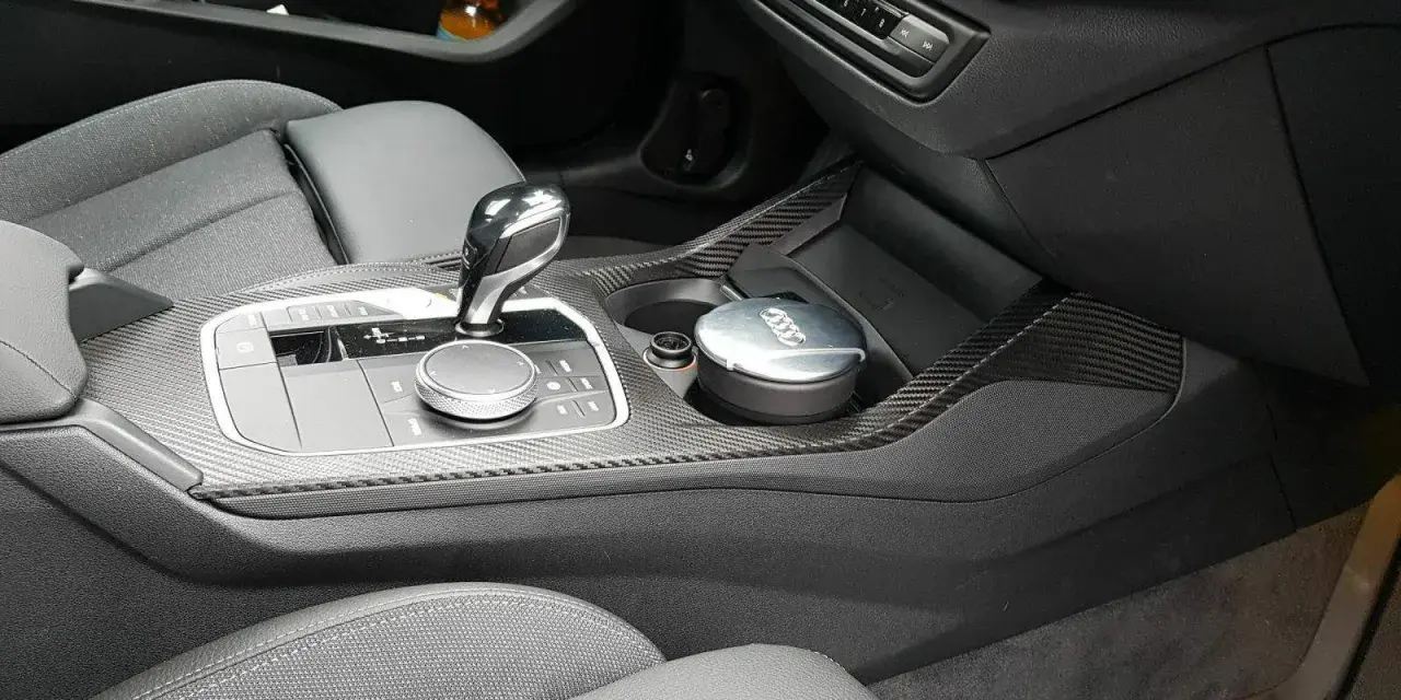 Detailaufnahme des Innenraums eines Autos mit Carbon-Akzenten. Ein Aschenbecher mit Audi-Logo steht im Getränkehalter. So reinigt man klavierlack auto.