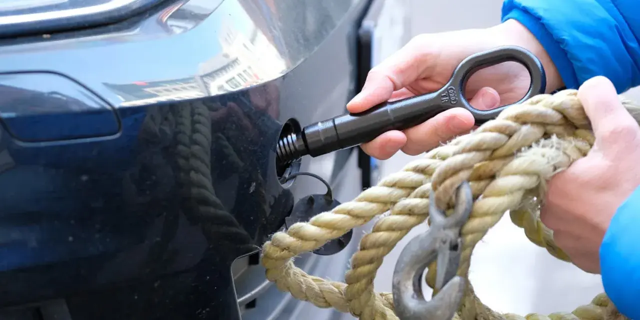 Eine Hand schraubt den Abschlepphaken in das Auto, während eine andere Hand ein dickes Seil mit Haken hält.
