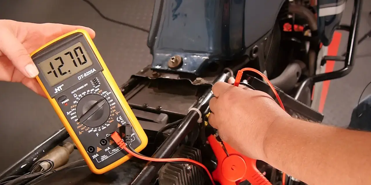Mit einem Multimeter die Lichtmaschine messen: Ein Mann hält das gelbe Gerät, das -12.70 Volt anzeigt, während die Prüfspitzen an der Motorradverkabelung anliegen.