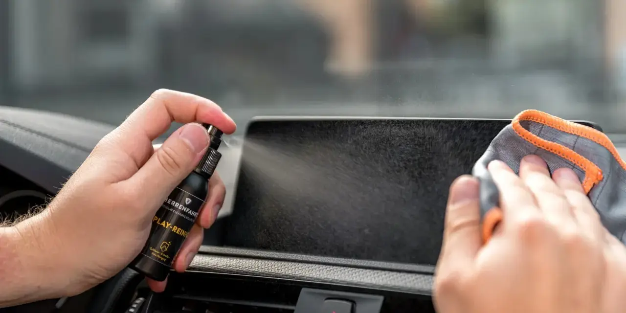 Mit einem Spray wird das Auto Display gereinigt. Eine Hand hält die Sprühflasche, die andere wischt mit einem Tuch über den Bildschirm.