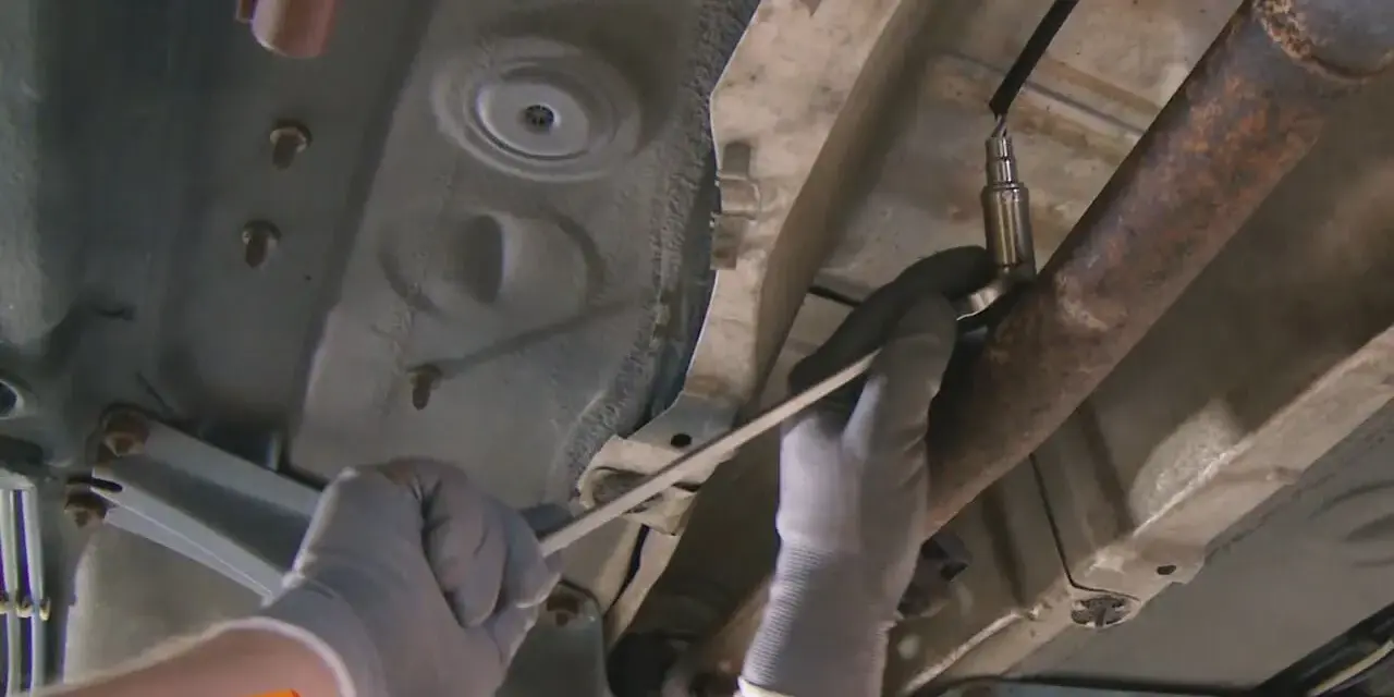 Ein Mechaniker löst die Lambdasonde am Auspuffrohr. Sie sitzt im vorderen Teil des Rohrs, nahe am Motor.