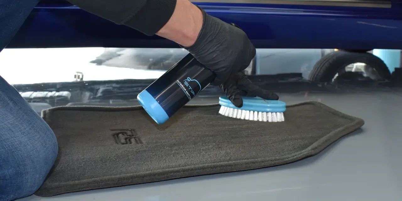 Person mit Handschuhen sprüht Reiniger auf einen grauen Autoteppich und schrubbt ihn mit einer Bürste, um ihn zu reinigen.