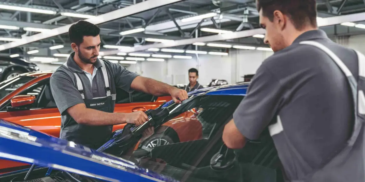 Zwei Mechaniker führen einen BMW Fahrzeugcheck durch. Einer entfernt die Windschutzscheibe, der andere sichert sie.