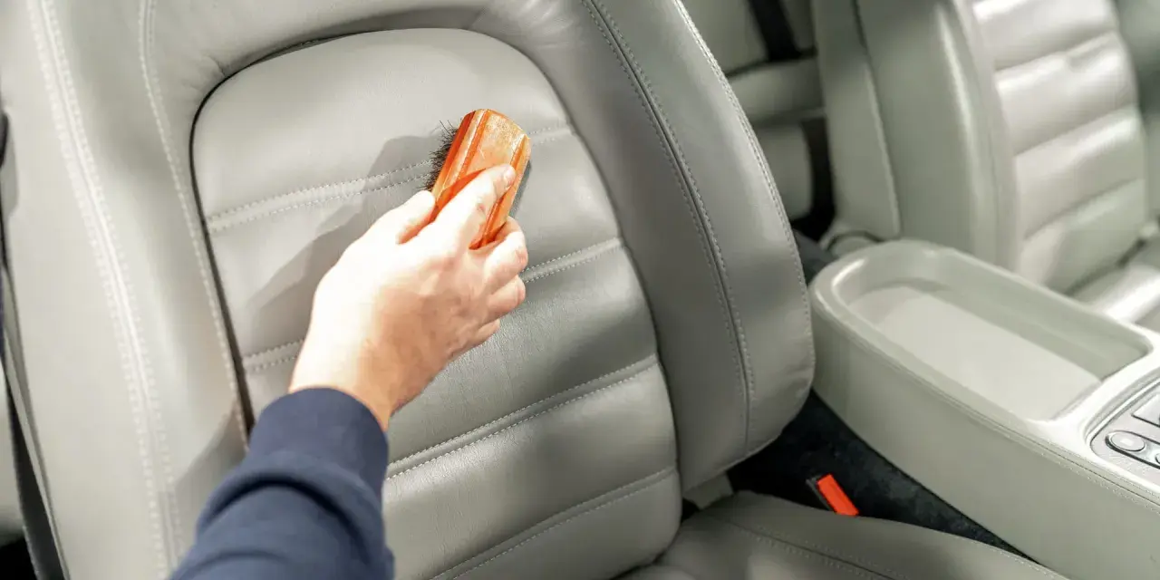 Hand reinigt grauen Leder-Autositz mit Bürste, um Wasserflecken zu entfernen.