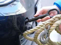 Eine Hand schraubt den Abschlepphaken in das Auto, während eine andere Hand ein dickes Seil mit Haken hält.