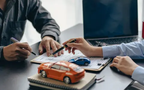 Zwei Personen unterschreiben einen Vertrag, während Modellautos und ein Laptop auf dem Tisch liegen. Dies symbolisiert den Kauf eines Autos mit Herstellergarantie.