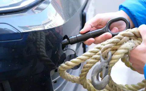 Eine Hand schraubt den Abschlepphaken in das Auto, während eine andere Hand ein dickes Seil mit Haken hält.