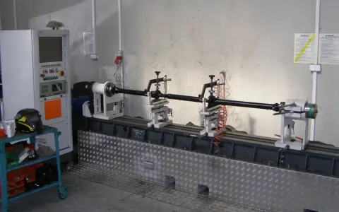 Kardanwelle wuchten auf einer speziellen Maschine. Die Welle ist auf Rollen gelagert, daneben eine Steuereinheit und ein Schweißhelm.