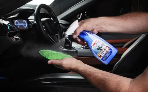 Mann sprüht Reinigungsmittel auf ein Mikrofasertuch, um Kunststoff im Auto zu reinigen.