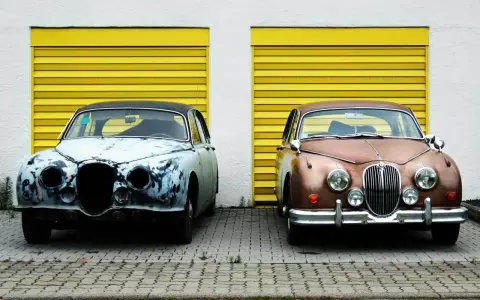 Zwei Oldtimer vor gelben Garagentoren. Einer ist rostig, der andere glänzt. Die **Oldtimer Lackieren Kosten** variieren stark.