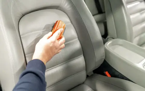 Hand reinigt grauen Leder-Autositz mit Bürste, um Wasserflecken zu entfernen.