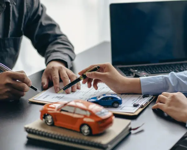 Zwei Personen unterschreiben einen Vertrag, während Modellautos und ein Laptop auf dem Tisch liegen. Dies symbolisiert den Kauf eines Autos mit Herstellergarantie.
