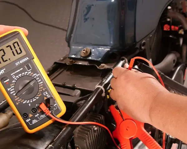 Mit einem Multimeter die Lichtmaschine messen: Ein Mann hält das gelbe Gerät, das -12.70 Volt anzeigt, während die Prüfspitzen an der Motorradverkabelung anliegen.