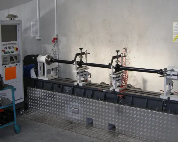 Kardanwelle wuchten auf einer speziellen Maschine. Die Welle ist auf Rollen gelagert, daneben eine Steuereinheit und ein Schweißhelm.