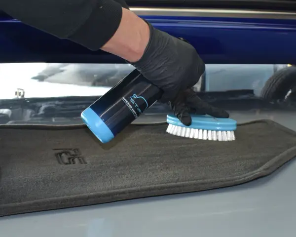 Person mit Handschuhen sprüht Reiniger auf einen grauen Autoteppich und schrubbt ihn mit einer Bürste, um ihn zu reinigen.