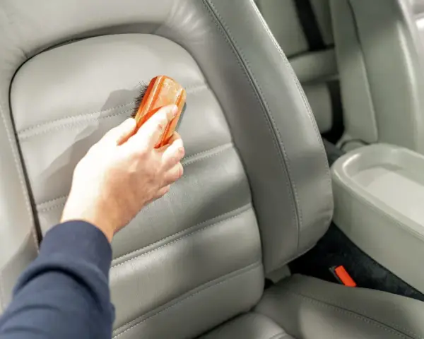 Hand reinigt grauen Leder-Autositz mit Bürste, um Wasserflecken zu entfernen.