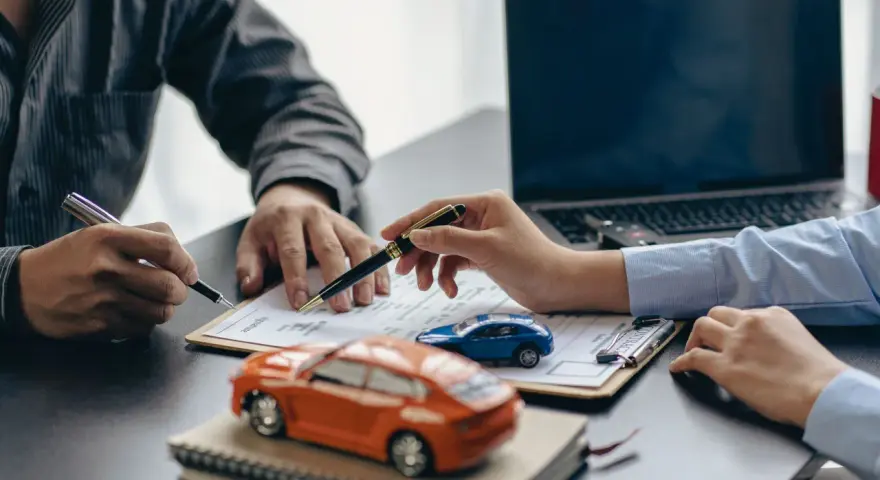 Zwei Personen unterschreiben einen Vertrag, während Modellautos und ein Laptop auf dem Tisch liegen. Dies symbolisiert den Kauf eines Autos mit Herstellergarantie.