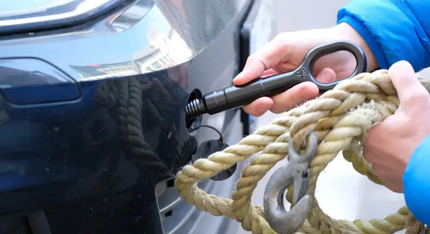 Eine Hand schraubt den Abschlepphaken in das Auto, während eine andere Hand ein dickes Seil mit Haken hält.