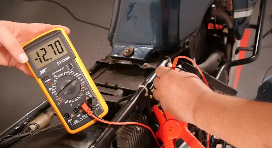 Mit einem Multimeter die Lichtmaschine messen: Ein Mann hält das gelbe Gerät, das -12.70 Volt anzeigt, während die Prüfspitzen an der Motorradverkabelung anliegen.