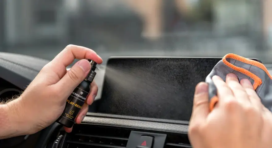 Mit einem Spray wird das Auto Display gereinigt. Eine Hand hält die Sprühflasche, die andere wischt mit einem Tuch über den Bildschirm.