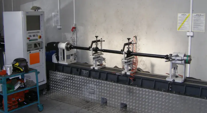 Kardanwelle wuchten auf einer speziellen Maschine. Die Welle ist auf Rollen gelagert, daneben eine Steuereinheit und ein Schweißhelm.