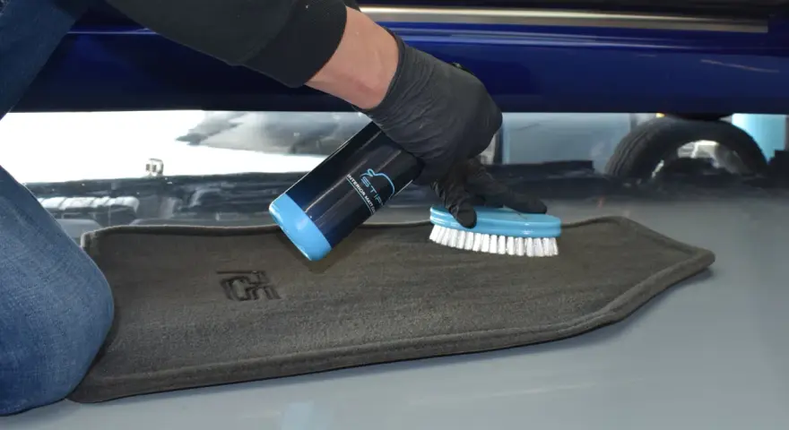 Person mit Handschuhen sprüht Reiniger auf einen grauen Autoteppich und schrubbt ihn mit einer Bürste, um ihn zu reinigen.