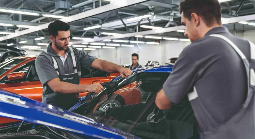 Zwei Mechaniker führen einen BMW Fahrzeugcheck durch. Einer entfernt die Windschutzscheibe, der andere sichert sie.