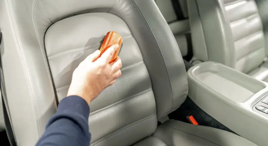 Hand reinigt grauen Leder-Autositz mit Bürste, um Wasserflecken zu entfernen.