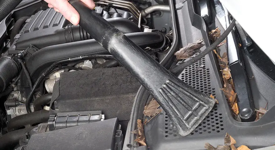 Hand saugt Laub und Schmutz aus dem Motorraum, um Feuchtigkeit im Auto vorzubeugen.
