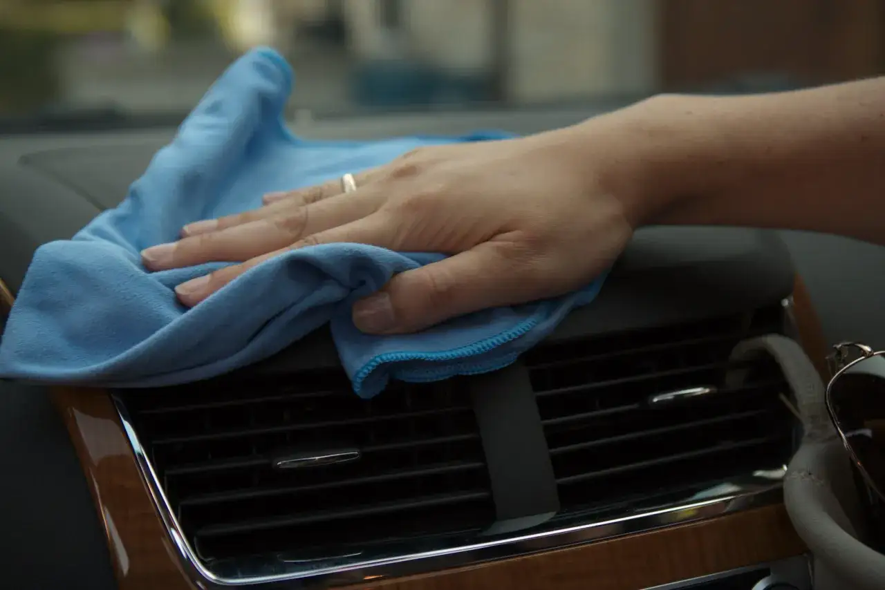 Hand wischt mit blauem Tuch &uuml;ber L&uuml;ftungsschlitze im Auto. So l&auml;sst sich Kunststoff im Auto reinigen und Staub entfernen.