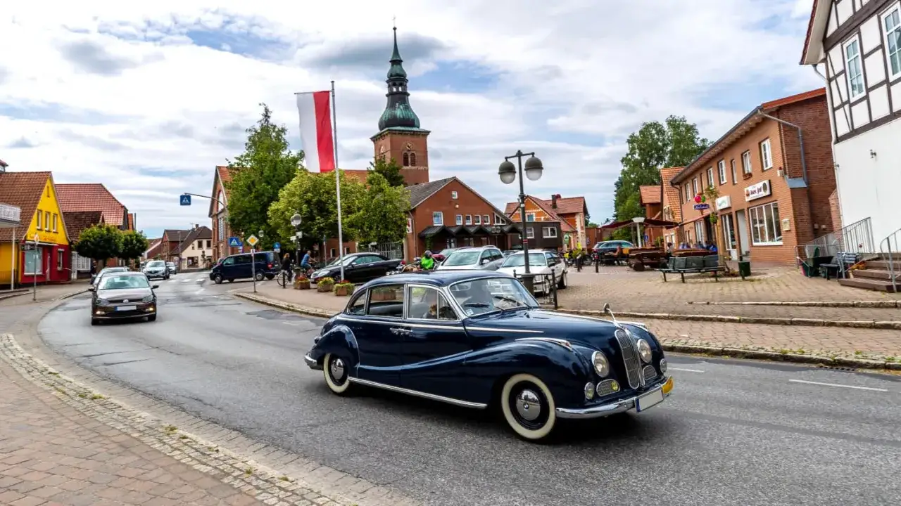 Ein glänzender Oldtimer fährt durch eine malerische deutsche Stadt. Die Kirche im Hintergrund und die Gebäude deuten auf eine Umweltzone hin, in der solche Klassiker willkommen sind.