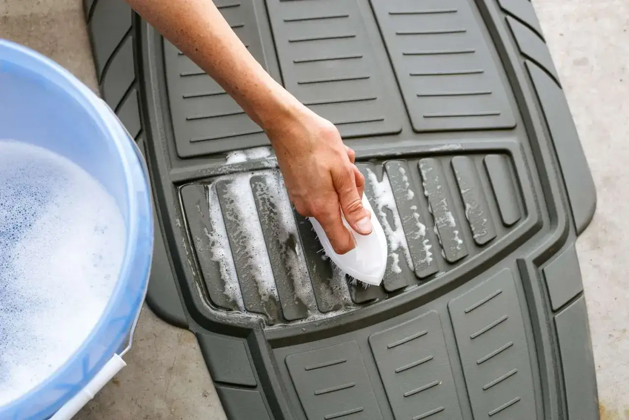 Hand schrubbt mit B&uuml;rste einen schmutzigen Autoteppich. Daneben steht ein Eimer mit Seifenwasser.