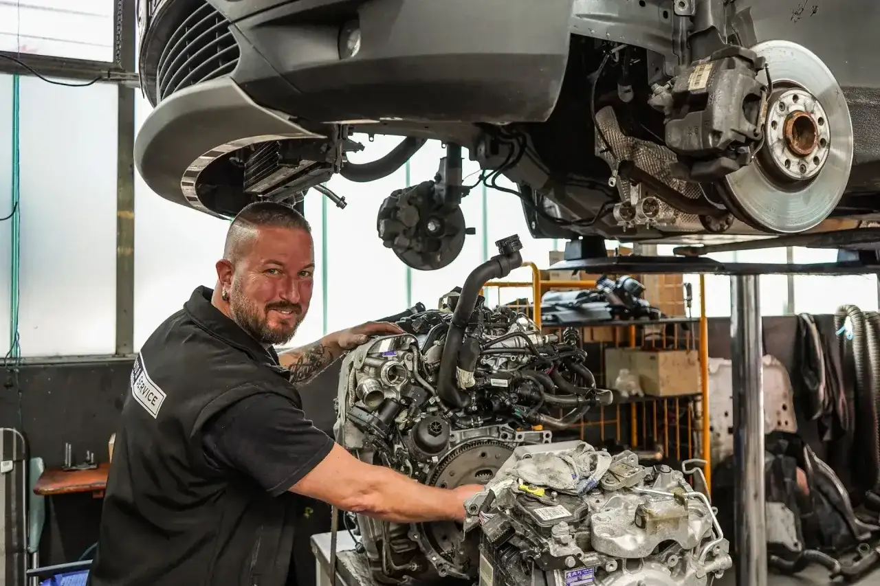 Mechaniker mit einem Motor im Vordergrund. Hier kann man seine pitstop teile selber mitbringen und sparen.