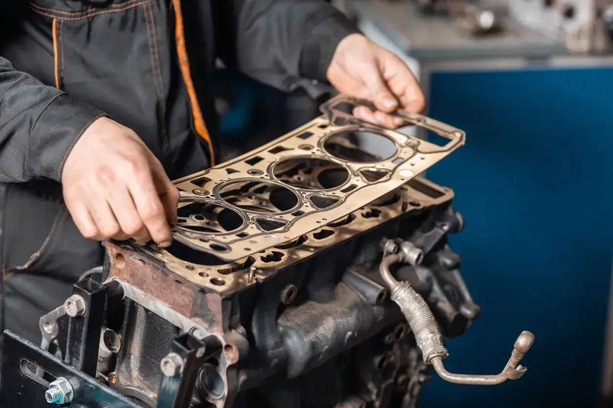 Mechaniker hält Zylinderkopfdichtung über Motorblock. Ein Motorschaden kann hohe Kosten verursachen, aber eine Reparatur ist oft möglich.