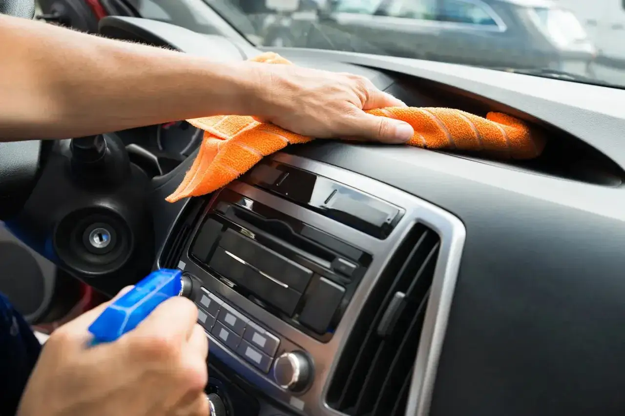 Hand spr&uuml;ht Reinigungsmittel auf das Armaturenbrett, w&auml;hrend ein orangefarbenes Tuch Staub wischt. Das Armaturenbrett wird aufbereitet.