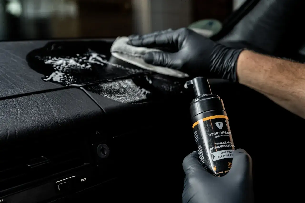 Mit schwarzer Handschuh und Reinigungsschaum das Armaturenbrett im Auto reinigen.