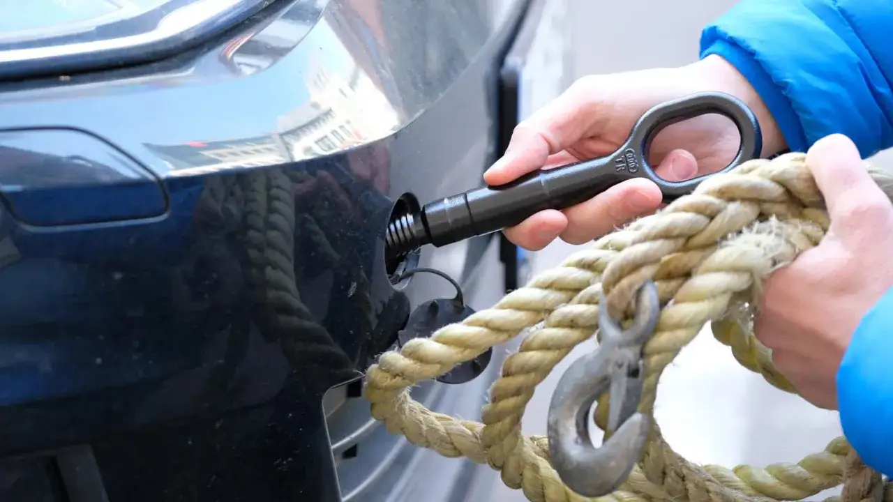 Eine Hand schraubt den Abschlepphaken in das Auto. Eine dicke Seil mit Haken liegt bereit zum Abschleppen.
