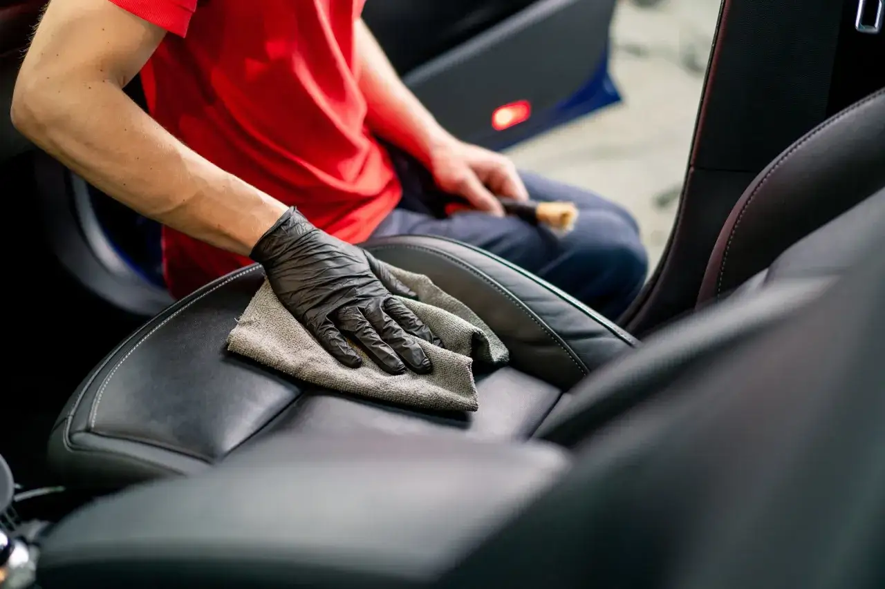 Person mit Handschuh wischt mit Tuch über schwarzes Leder im Auto. Sorgfältiges leder reinigen für ein makelloses Interieur.