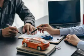 Zwei Personen unterschreiben einen Vertrag, während Modellautos und ein Laptop auf dem Tisch liegen. Dies symbolisiert den Kauf eines Autos mit Herstellergarantie.