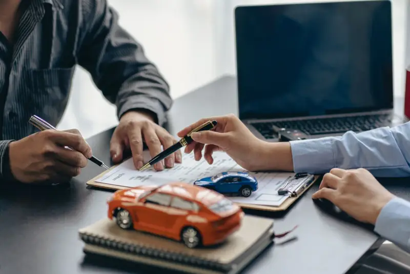Zwei Personen unterschreiben einen Vertrag, während Modellautos und ein Laptop auf dem Tisch liegen. Dies symbolisiert den Kauf eines Autos mit Herstellergarantie.