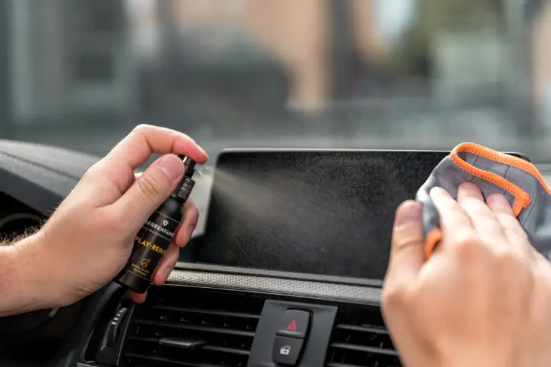 Mit einem Spray wird das Auto Display gereinigt. Eine Hand hält die Sprühflasche, die andere wischt mit einem Tuch über den Bildschirm.